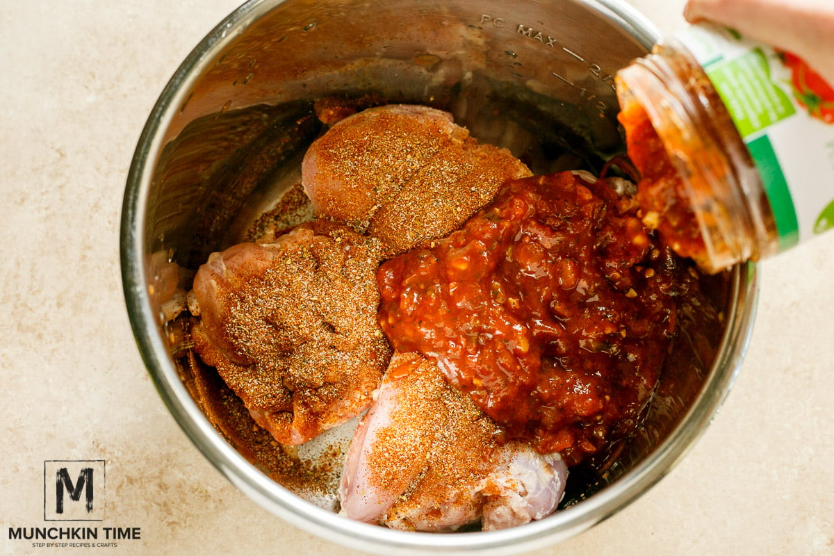 Raw chicken thighs inside crockpot, with homemade taco seasoning and salsa sauce pouring out of the jar.