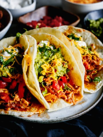 3 Juicy Chicken Tacos on a grey plate with juicy shredded chicken, guacamole, lettuce, cheese and sour cream.