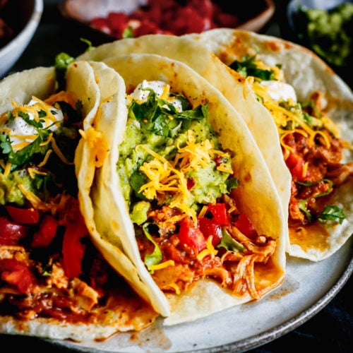 3 Juicy Chicken Tacos on a grey plate with juicy shredded chicken, guacamole, lettuce, cheese and sour cream.