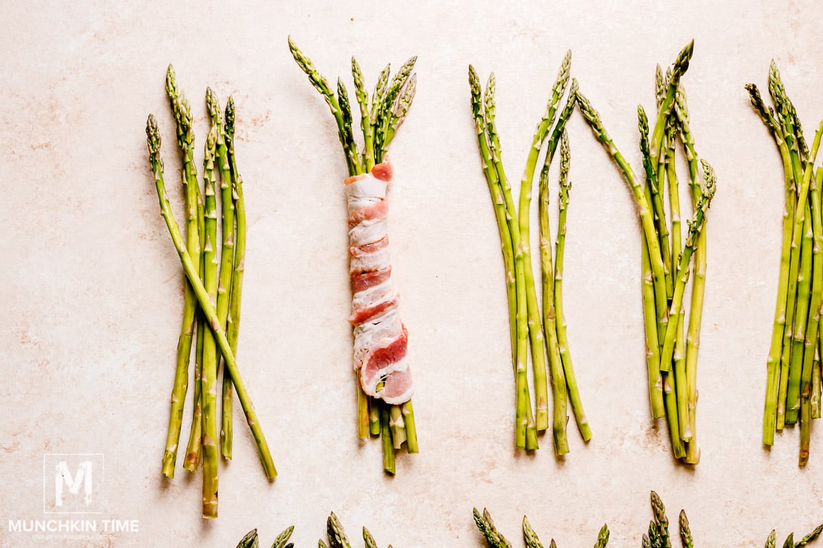 Asparagus bundles on a surface, one bundle is wrapped in bacon. 