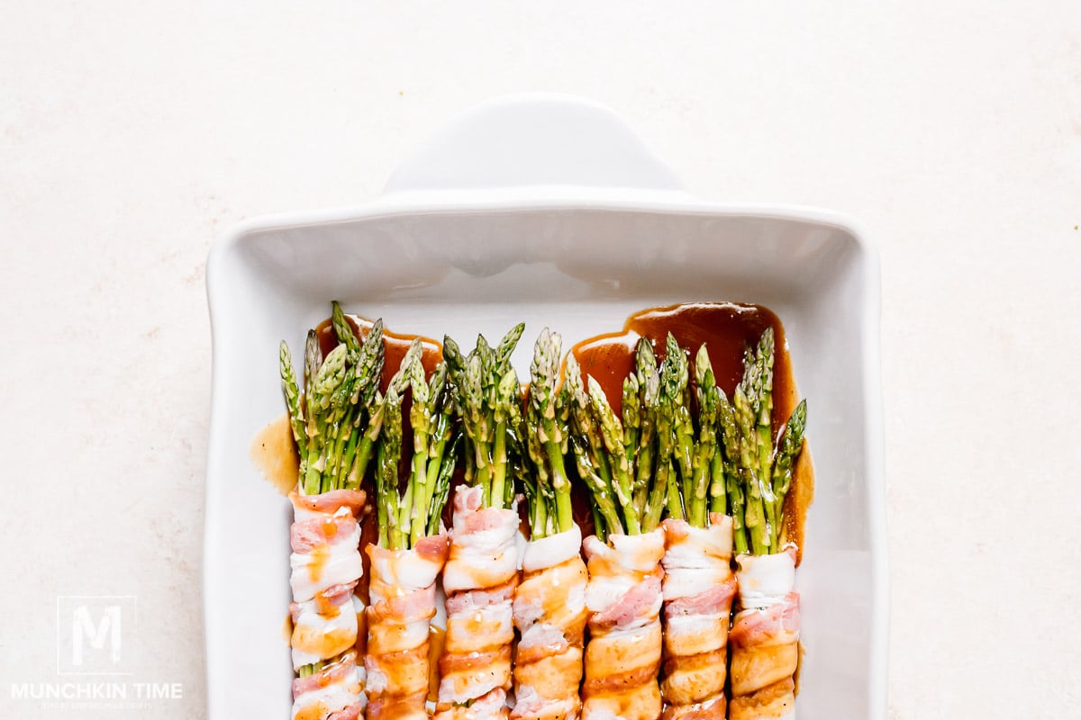 Warm sauce is poured over the asparagus bundles wrapped in bacon, ready to go into the oven.