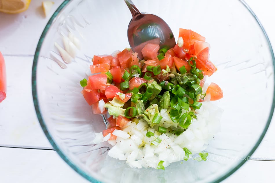 A mixing bowl of Bruschetta filling. Inside the bowl are chopped onion, cubed avocado, cubed tomatoes, salt, pepper and green onion. 