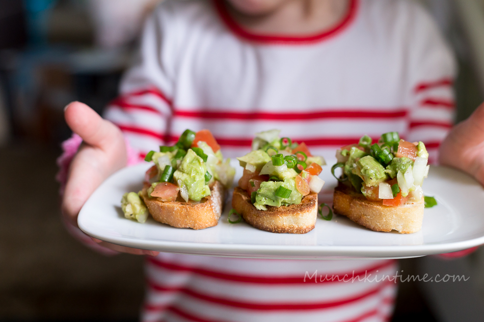 A little girl holding a plate with 3 Tomato Bruschetta garish with avocado and green onion.