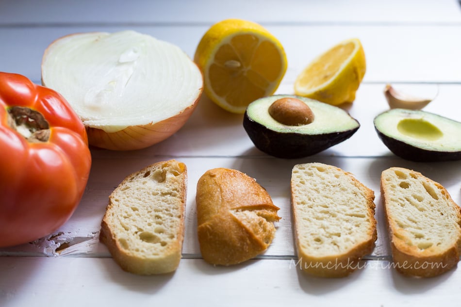 All of the ingredients for Tomato Avocado Bruschetta Recipe laying on the table, they are tomato, onion, lemon, avocado, garlic and sliced bread. 