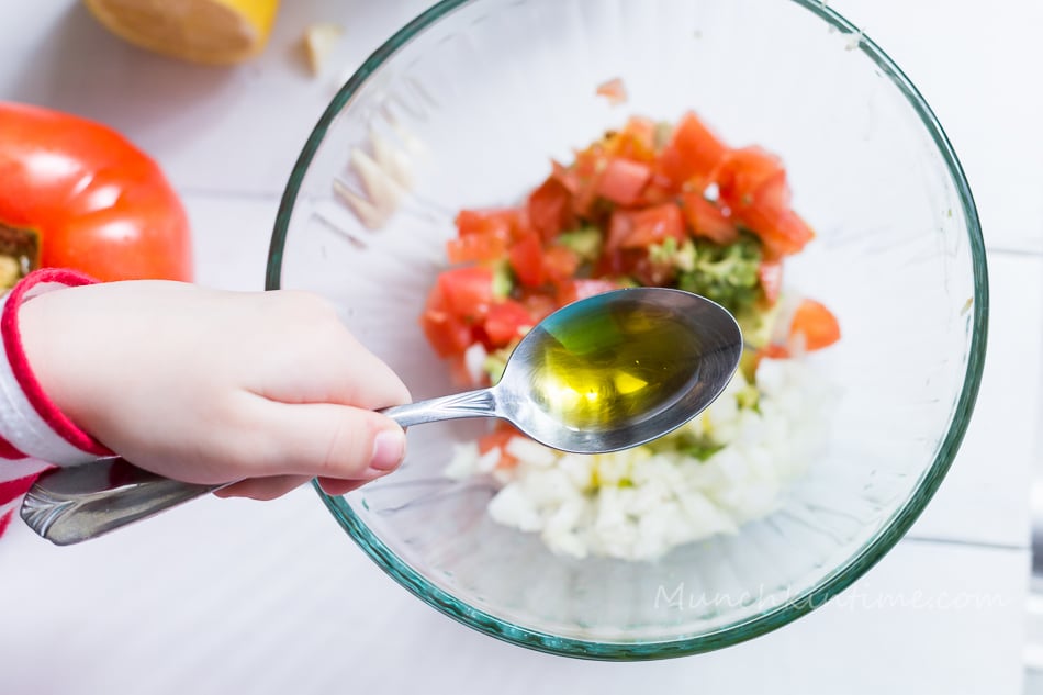 A hand holding a spoon of olive oil over the bruschetta filling. 