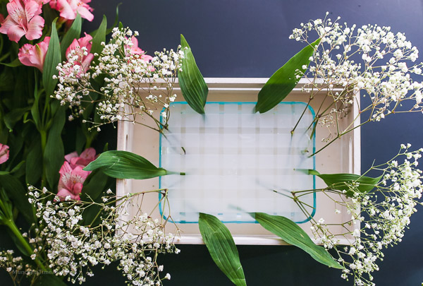 The glass container placed inside the wooden box and fresh flowers on the side to make flowers centerpiece.  First flower layer is added to the glass container. 