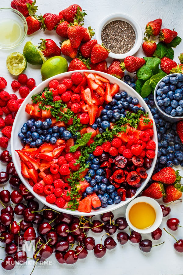 A large bowl filled with sliced strawberries, blueberries, raspberries, and cherries, surrounded by fresh fruit, mint, honey, lime, and chia seeds on a white surface.