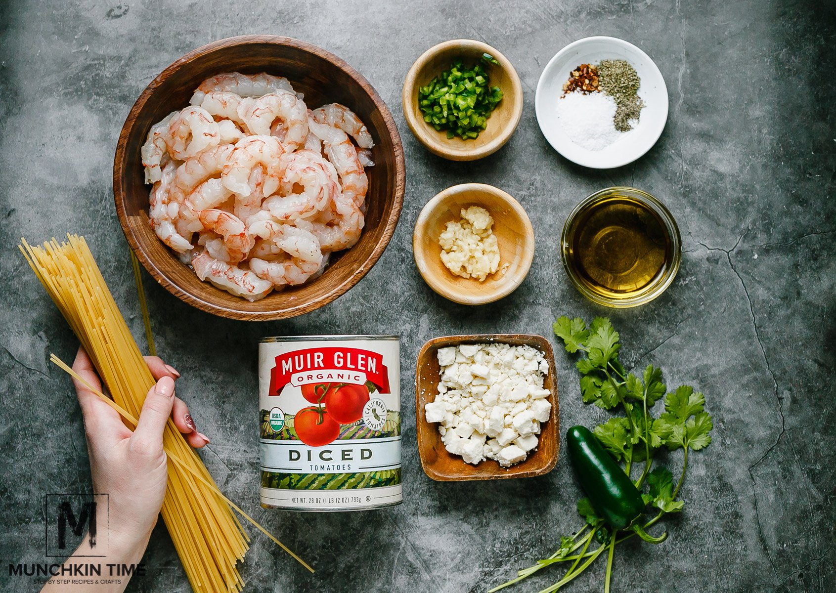 All ingredients on a table for Italian Shrimp Pasta. They are pasta, raw shrimp, tomatoes in a can, feta cheese, onion, garlic, seasonings, onion, jalapeño and greens.