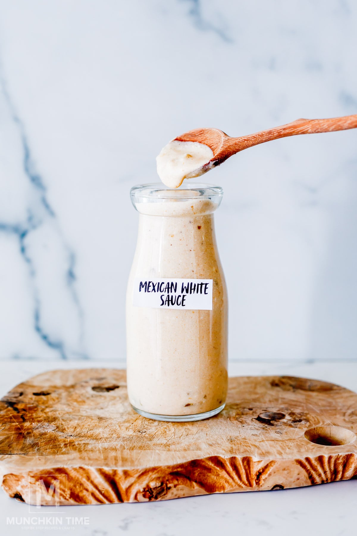A glass jar labeled "Mexican White Sauce" sits on a wooden board, with a wooden spoon holding some of the sauce above the jar.