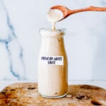 A glass bottle labeled "Mexican White Sauce" sits on a wooden board, while a wooden spoon drips sauce into the bottle.