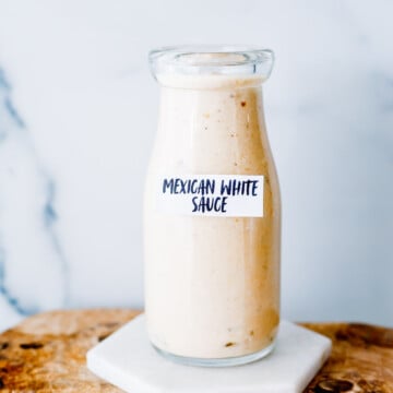 A glass bottle labeled "Mexican White Sauce" is filled with a creamy, light-colored sauce and placed on a marble coaster atop a wooden surface.