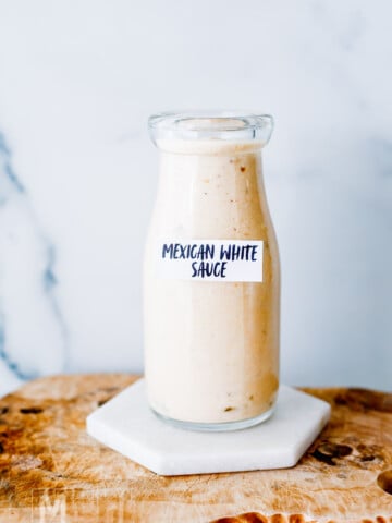 A glass bottle labeled "Mexican White Sauce" is filled with a creamy, light-colored sauce and placed on a marble coaster atop a wooden surface.