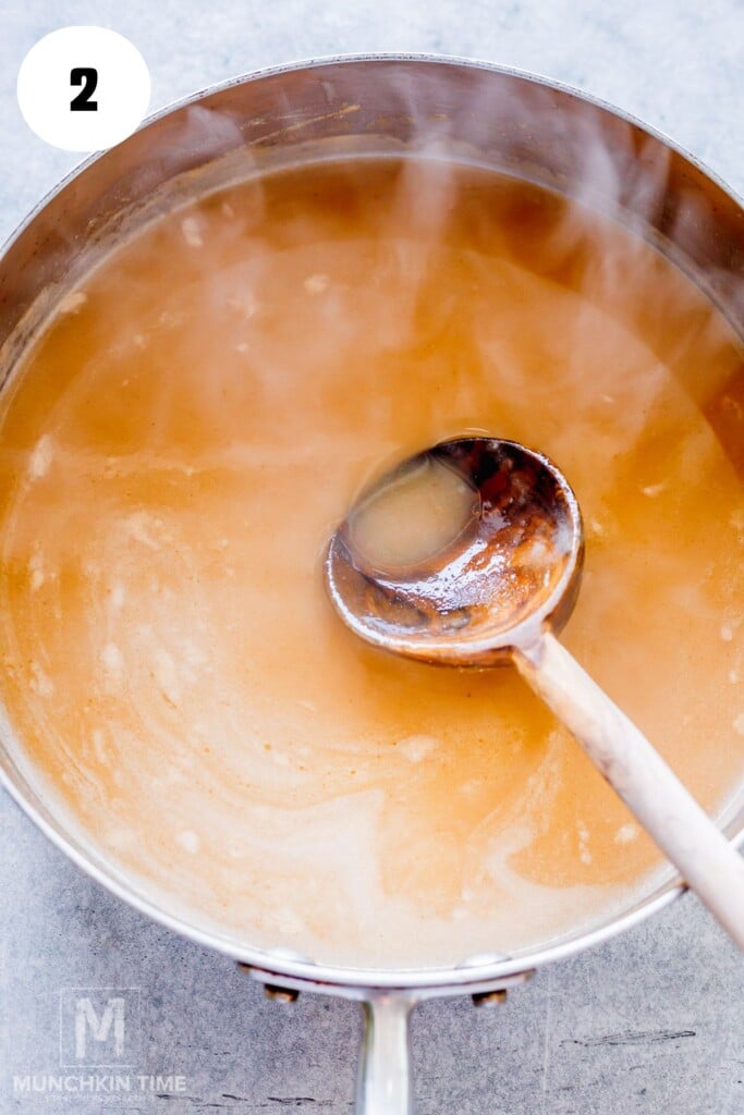A steaming pot of simmering brown broth being stirred with a wooden ladle, viewed from above.