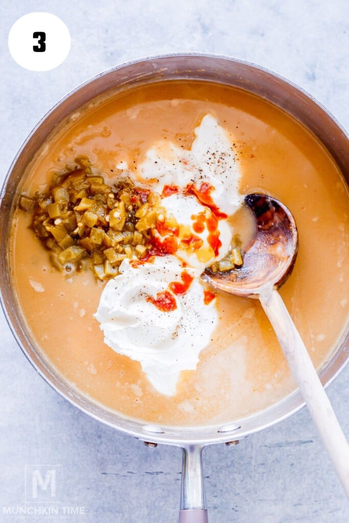 A pot of soup with sour cream, chopped green chilies, black pepper, and hot sauce added, being mixed with a wooden spoon.