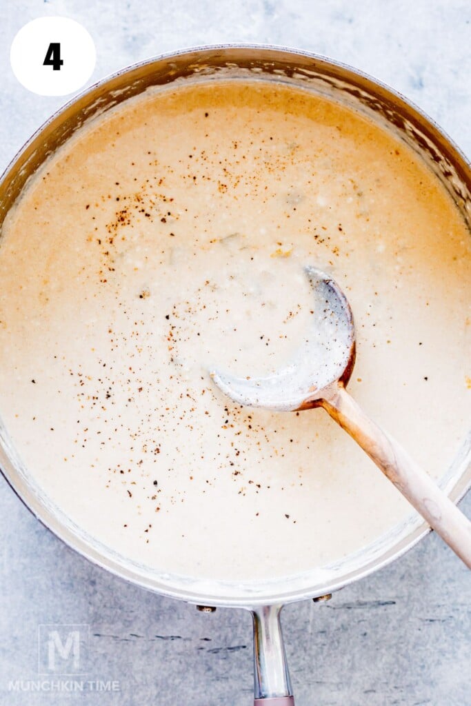 A saucepan filled with creamy sauce, sprinkled with black pepper, with a wooden spoon resting inside.