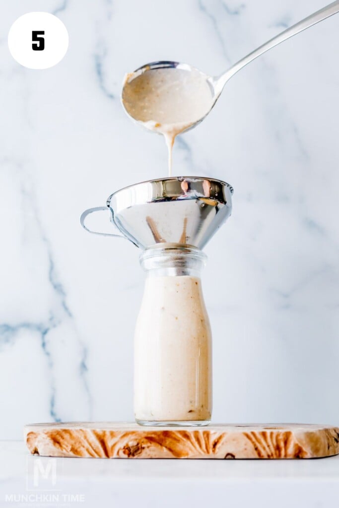 A creamy sauce is being poured from a ladle through a metal funnel into a glass bottle, which is placed on a wooden board.