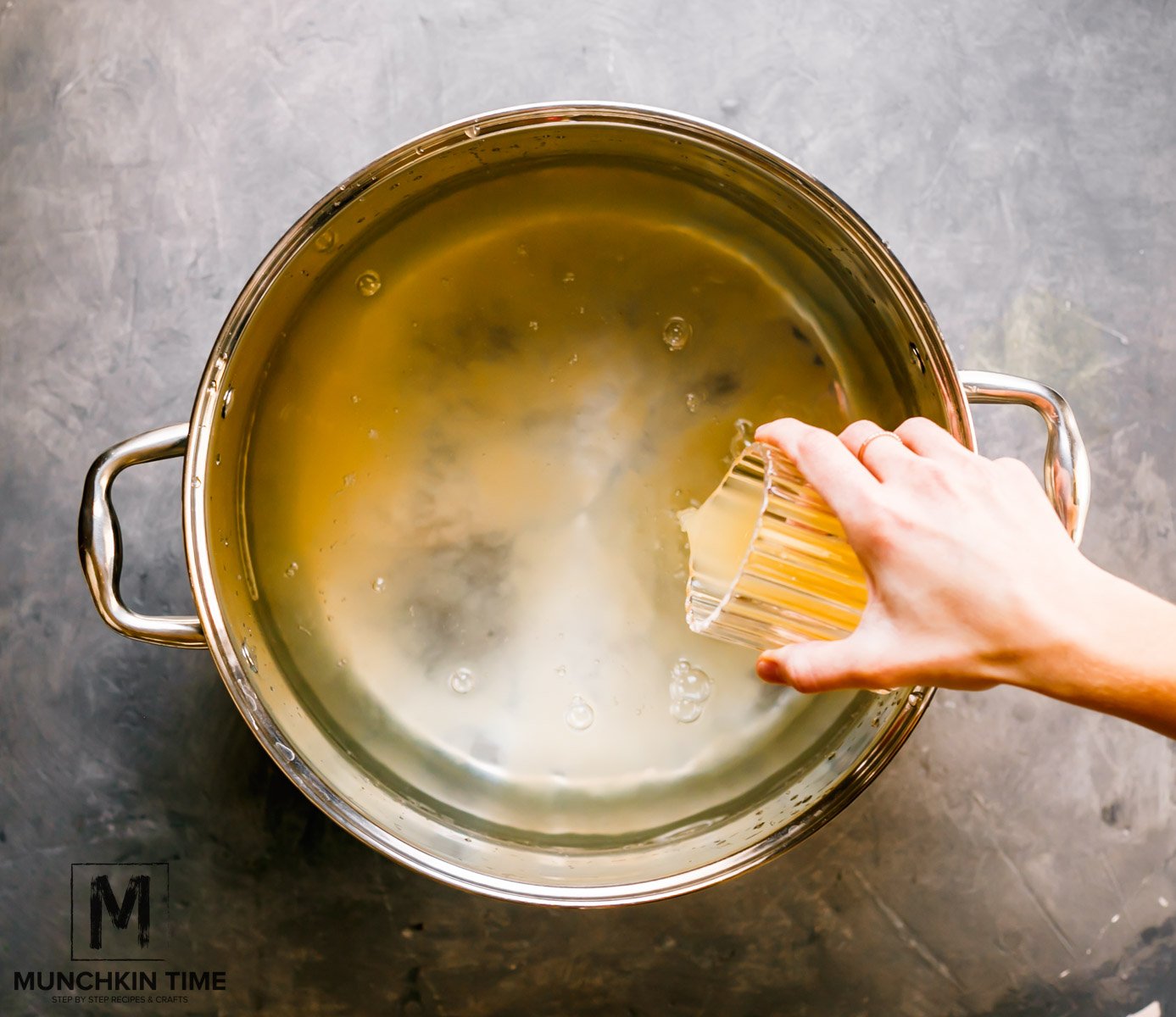 A hand pouring apple juice from a glass cup into a big pot filled with water.