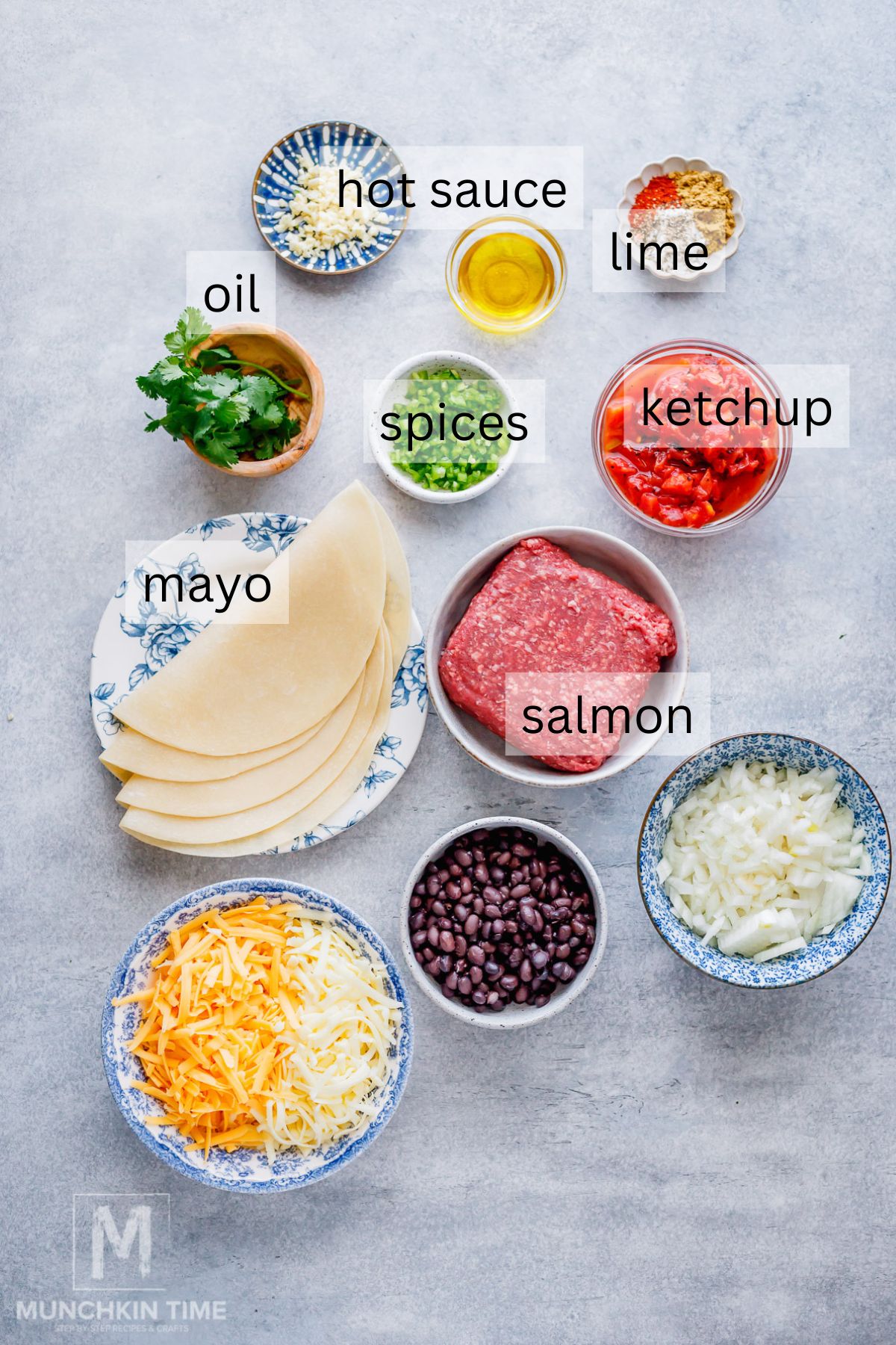 Assorted ingredients in bowls including shredded cheese, black beans, chopped onion, mayo, salmon, spices, oil, lime, hot sauce, ketchup, cilantro, garlic, and folded tortillas.