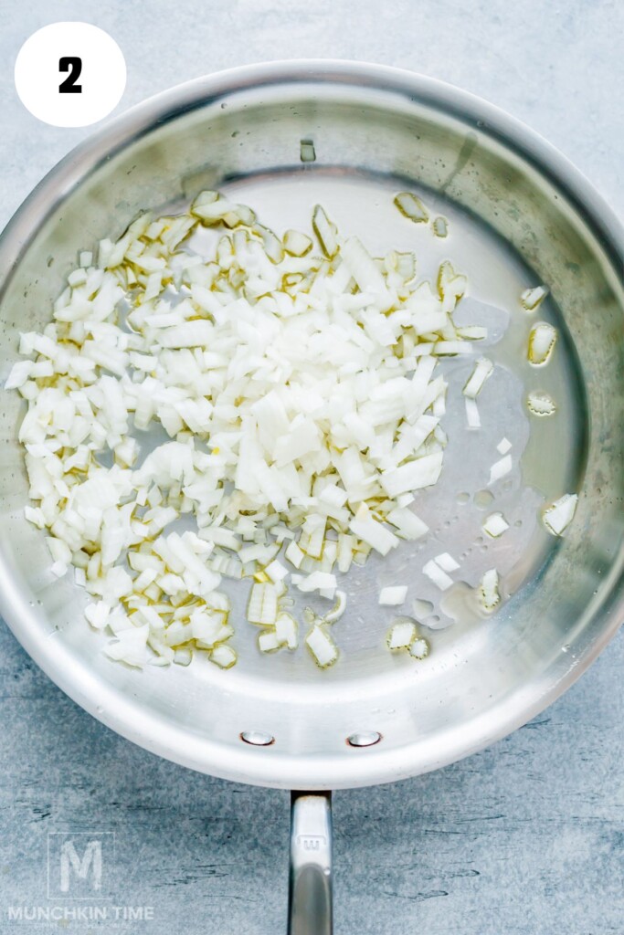 Diced onions cooking in oil in a stainless steel skillet on a light-colored surface, labeled with the number 2 in a white circle.
