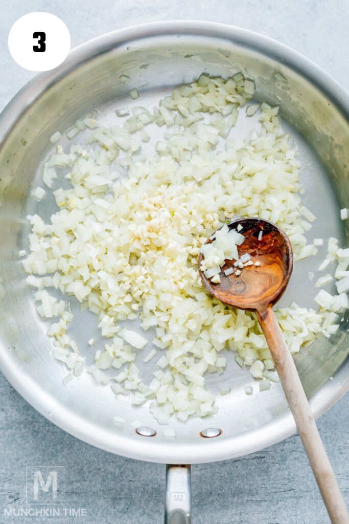 Chopped onions and garlic being sautéed in a stainless steel pan with a wooden spoon.