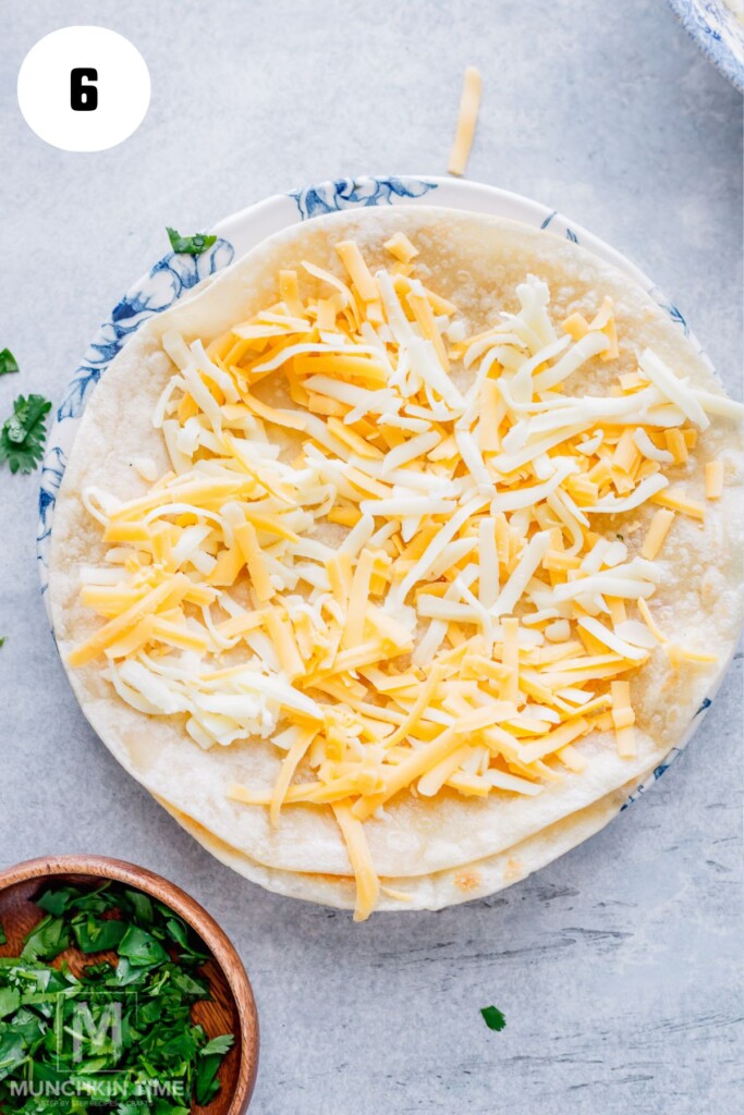 A tortilla on a plate topped with shredded yellow and white cheese, with a small bowl of chopped herbs nearby.