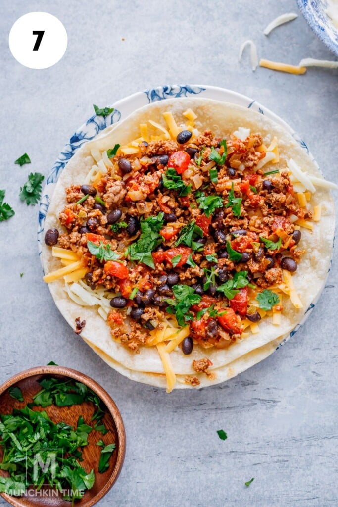 A tortilla on a plate topped with shredded cheese, ground meat, black beans, diced tomatoes, and chopped cilantro. A small bowl of cilantro is nearby.