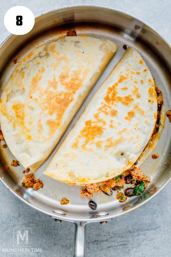 A stainless steel pan containing two folded, golden-brown quesadillas filled with a mixture of ground meat, black beans, and greens.