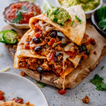 A stack of cheesy quesadillas filled with ground meat, black beans, and vegetables sits on a wooden board, topped with guacamole. Salsa, lime wedges, and jalapeño slices are nearby.