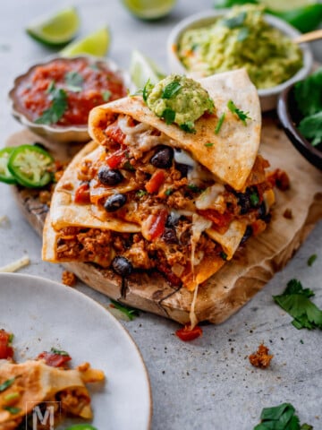 A stack of cheesy quesadillas filled with ground meat, black beans, and vegetables sits on a wooden board, topped with guacamole. Salsa, lime wedges, and jalapeño slices are nearby.