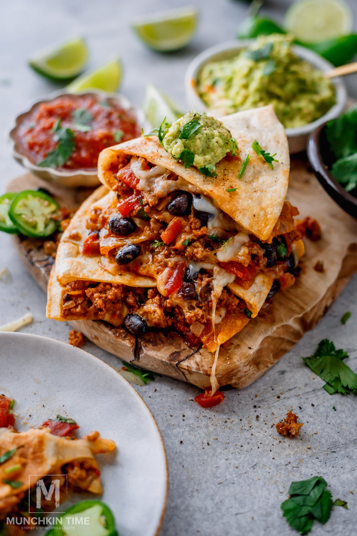 A stack of cheesy quesadillas filled with ground meat, black beans, and vegetables sits on a wooden board, topped with guacamole. Salsa, lime wedges, and jalapeño slices are nearby.