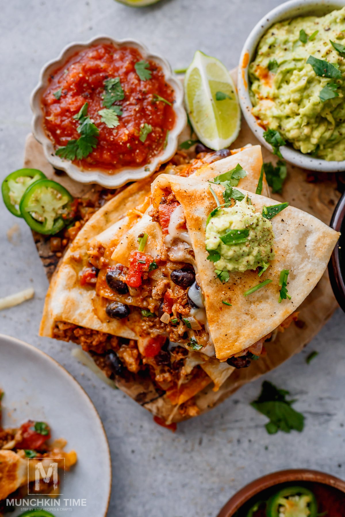 A stack of quesadilla slices filled with beans, meat, and vegetables, topped with guacamole and chopped cilantro, served with salsa and lime wedges.