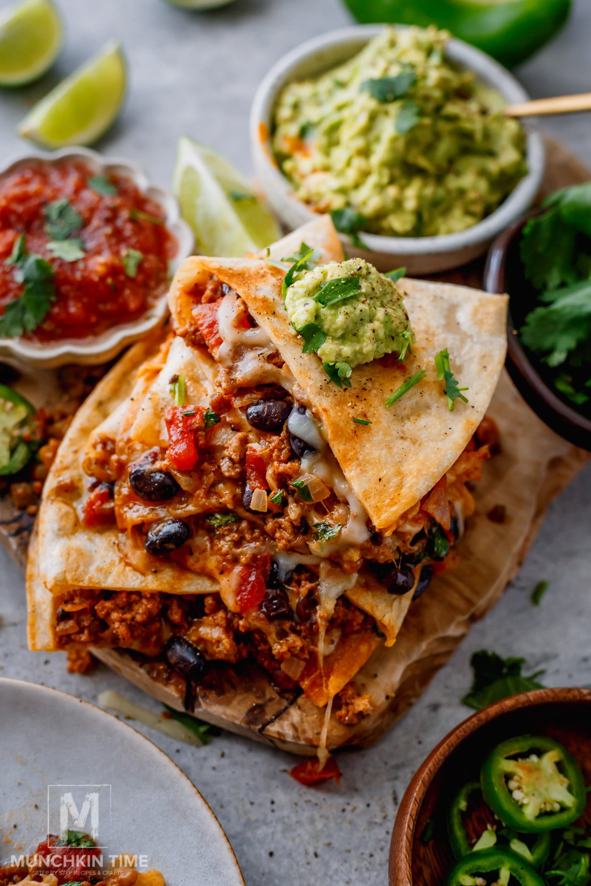 A stack of cheesy quesadillas filled with ground meat, black beans, and tomatoes, topped with guacamole, with salsa, guacamole, jalapeños, and lime wedges on the side.
