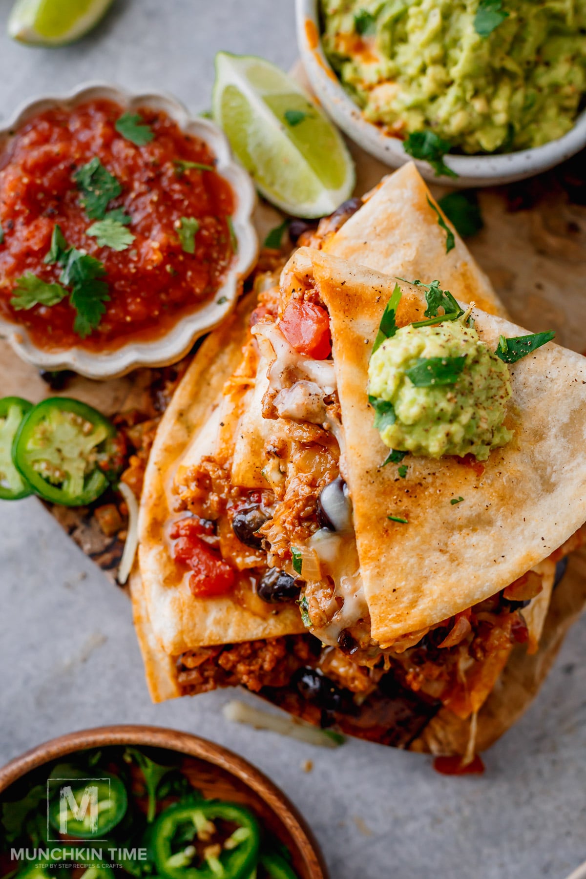 A stack of quesadillas filled with meat, beans, and cheese, topped with guacamole, with salsa, guacamole, lime wedges, and sliced jalapeños on the side.