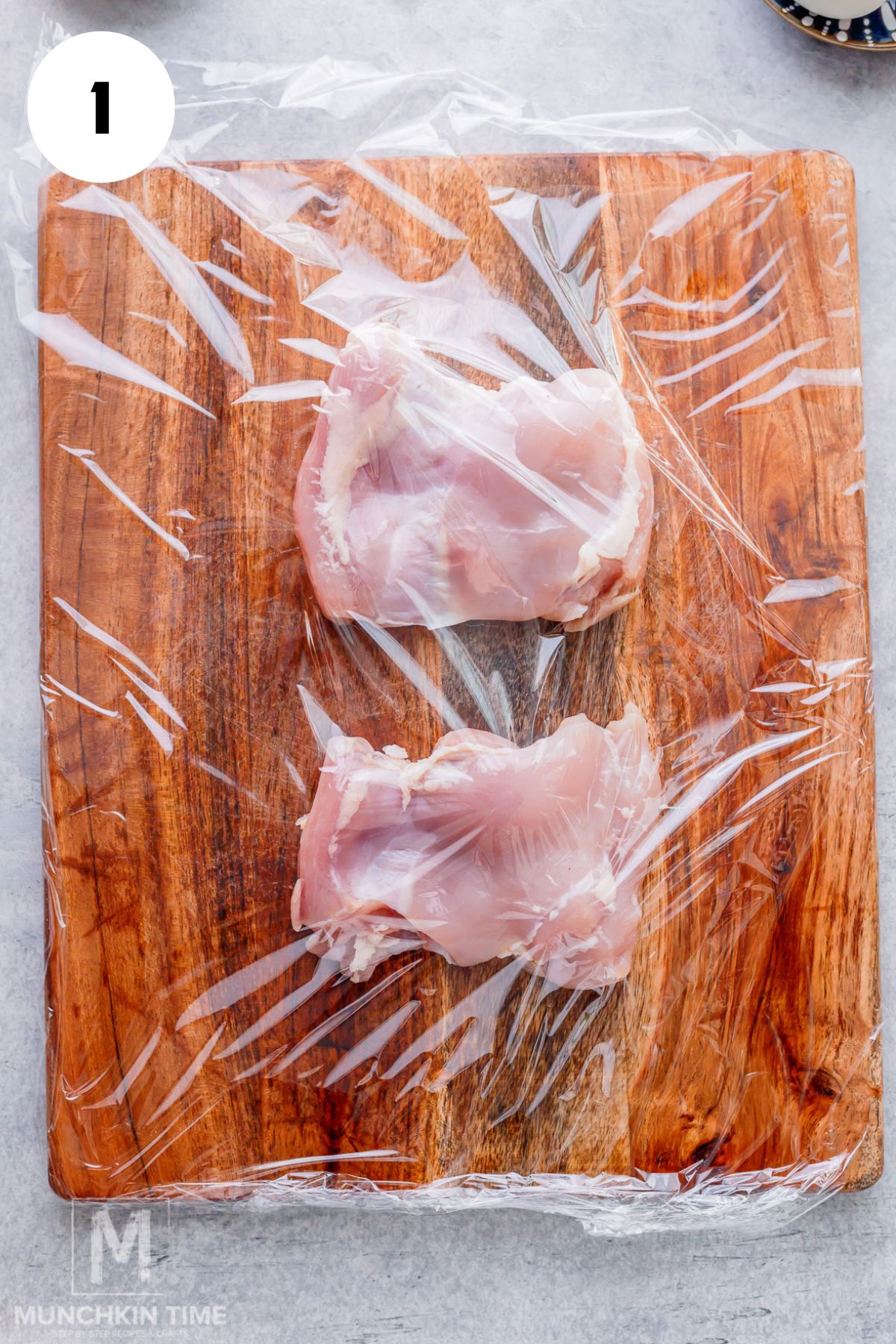two chicken thighs on a cutting board with plastic wrap on top, ready to be pounded.
