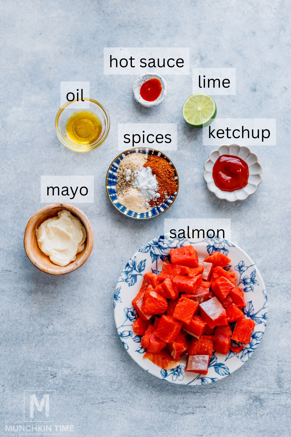 A plate of cubed salmon with small bowls containing mayonnaise, oil, spices, hot sauce, lime, and ketchup, all labeled on a light background.