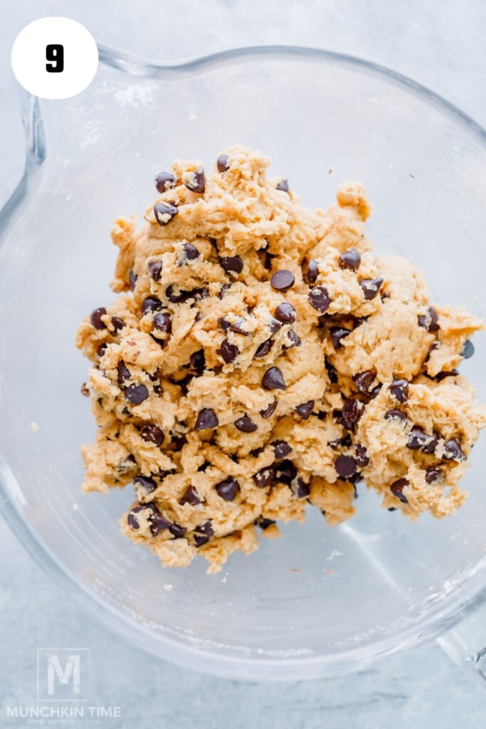 A glass mixing bowl filled with chocolate chip cookie dough sits on a light surface.