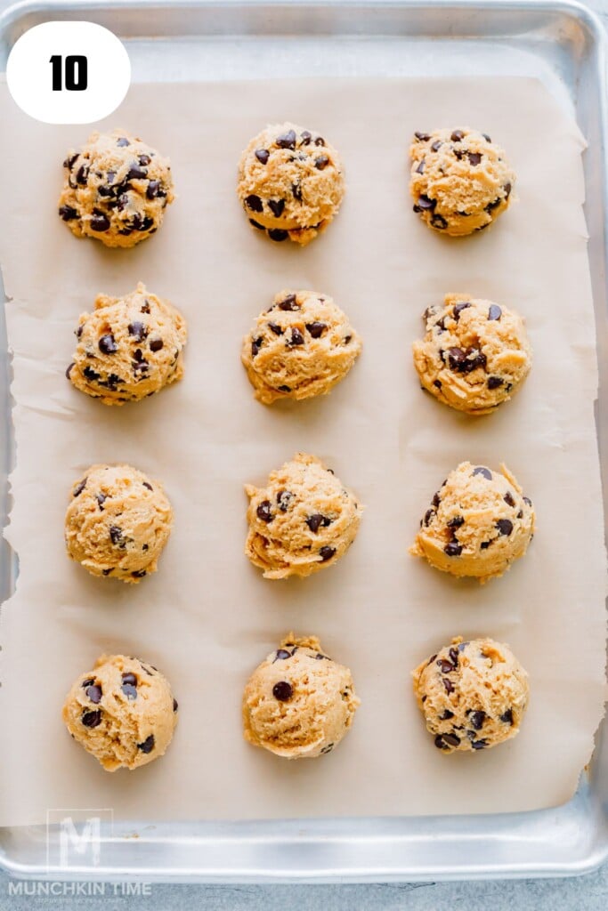 Twelve scoops of chocolate chip cookie dough arranged in rows on a parchment-lined baking sheet.