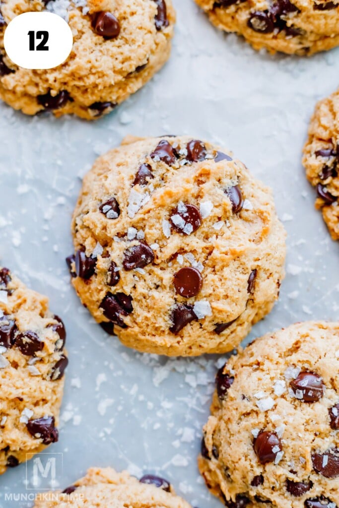 A close-up of sourdough chocolate chip cookies topped with flaky sea salt on a light, textured surface.