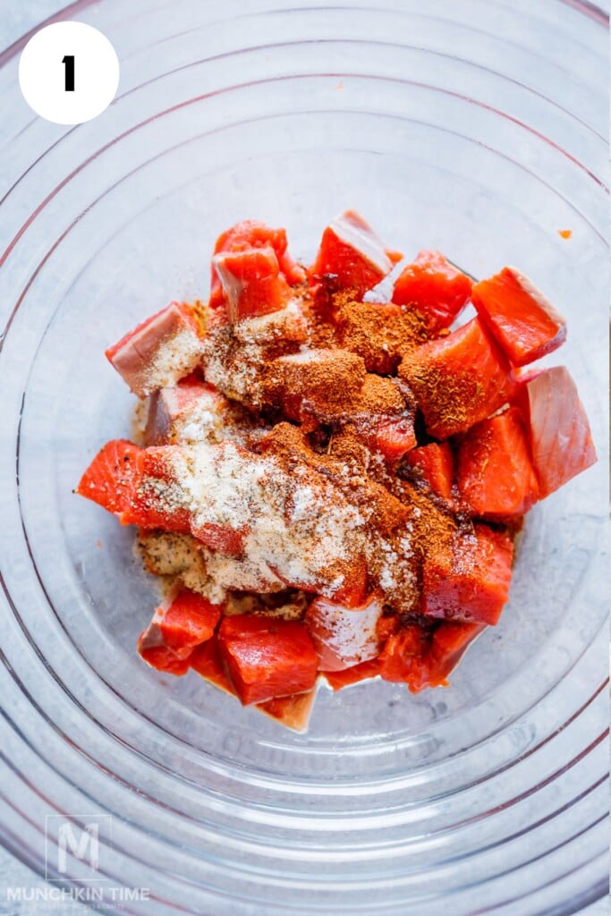 Cubed raw salmon in a glass bowl topped with various powdered spices, photographed from above.