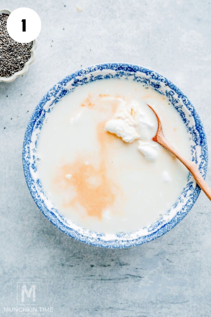 A blue patterned bowl filled with milk, yogurt, and vanilla, with a wooden spoon; chia seeds in a small dish nearby.