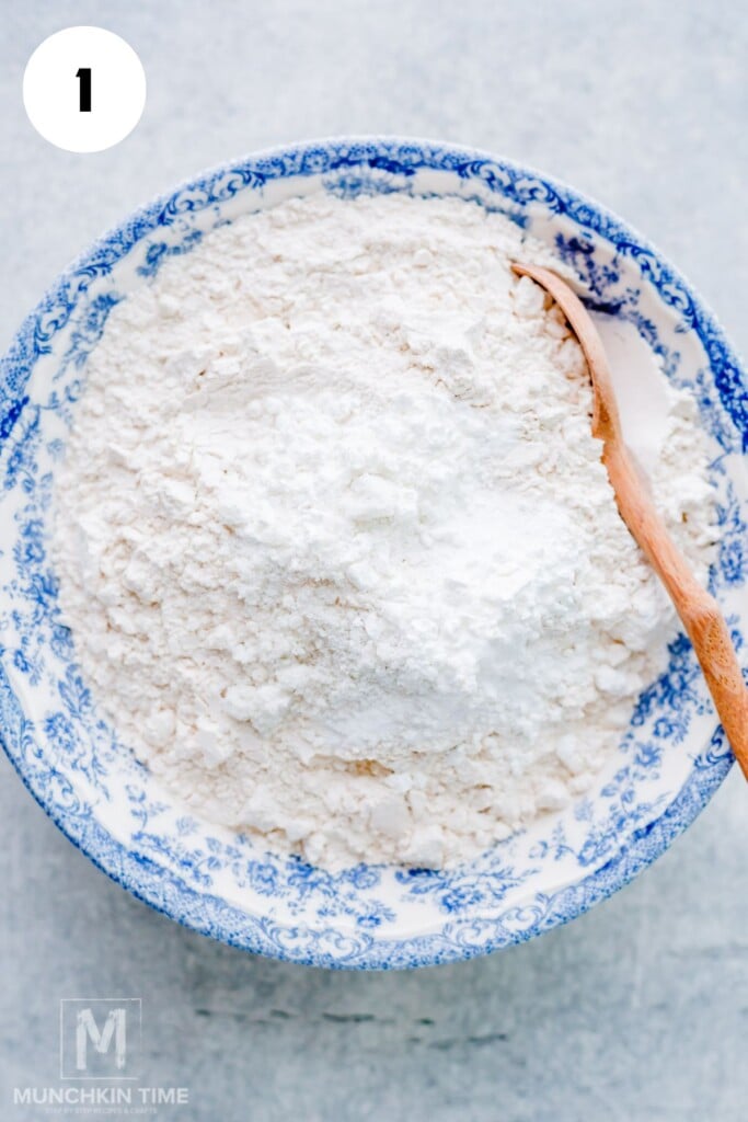 Blue and white bowl filled with flour mixture and a wooden spoon, viewed from above on a light surface.