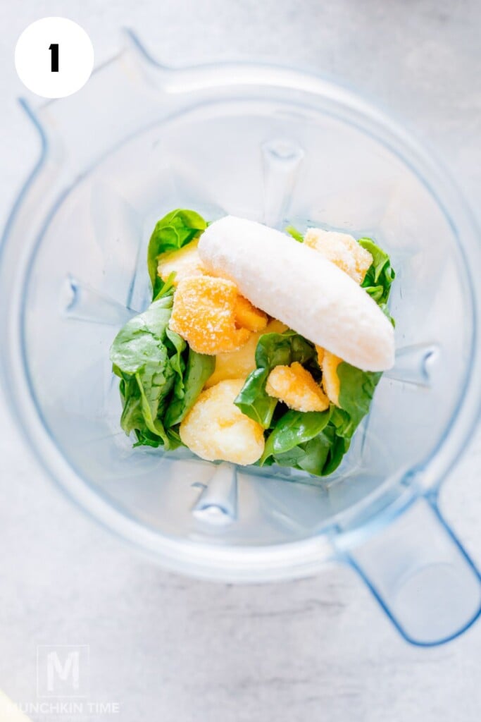 Overhead view of a blender containing spinach, a peeled banana, and pieces of frozen fruit, ready to be blended.