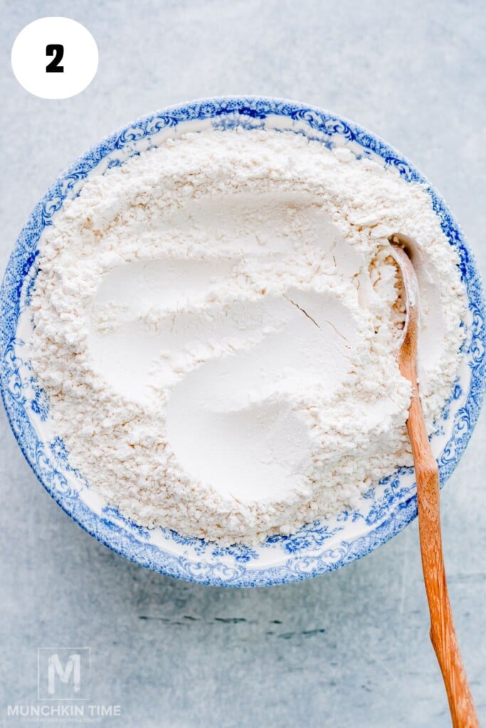 A blue and white bowl filled with flour and a wooden spoon, viewed from above, on a light surface.
