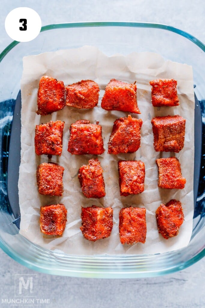 Fifteen pieces of seasoned salmon bites are arranged on parchment paper inside a glass baking dish.