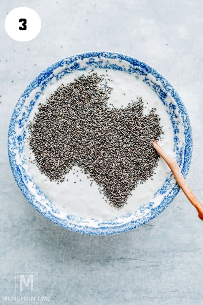 A blue and white bowl of yogurt is topped with chia seeds arranged in the shape of Australia, with a wooden spoon resting on the right side.