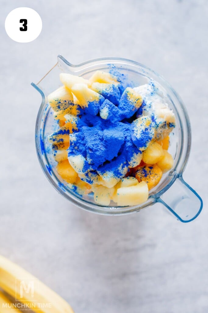 A blender filled with chopped bananas, pineapple chunks, yogurt, and bright blue powder, viewed from above.