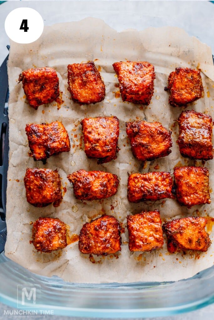 Fourteen pieces of seasoned, air fryer salmon bites arranged on parchment paper in a glass baking dish.