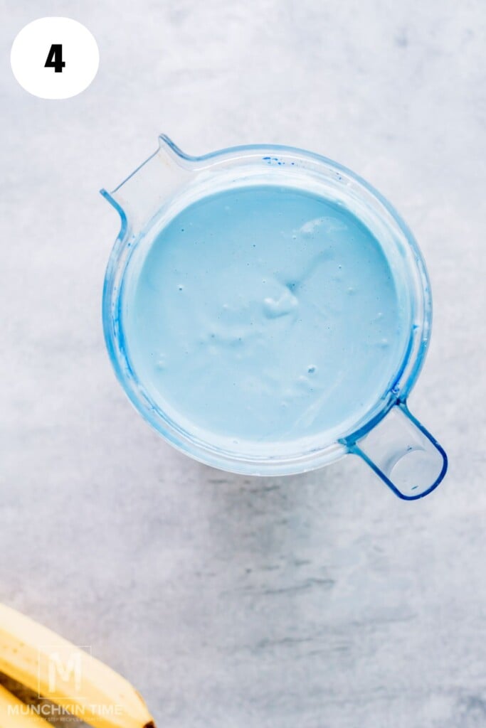 Top view of a blender containing a smooth, light blue mixture on a gray surface, with a banana partially visible in the bottom left corner.