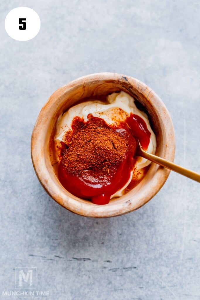 A wooden bowl containing mayonnaise, ketchup, and a pile of red seasoning powder, with a spoon inside.