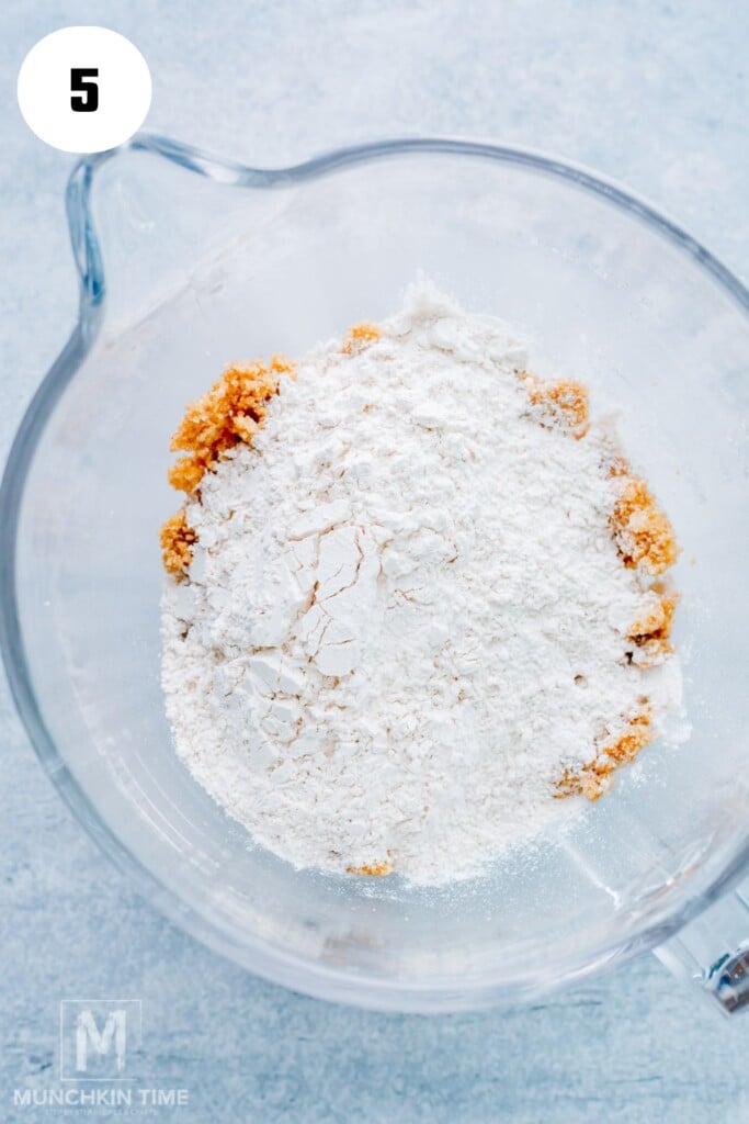 A glass mixing bowl containing white flour on top of a brown sugar mixture, viewed from above. Number 5 is in a white circle in the upper left corner.